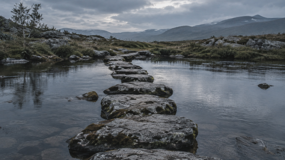 Worn stepping stones crossing calm water toward distant mountains, representing that generosity isn't the same as growth without real effort.